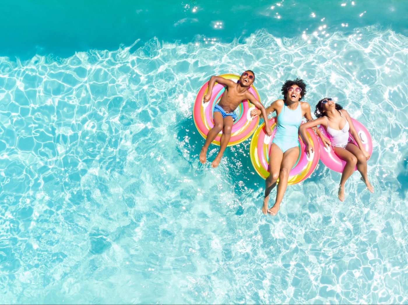 Three people—two children and an adult—float joyfully on colorful pool inflatables in a bright, clear blue swimming pool under the sun. ©wavebreakmedia