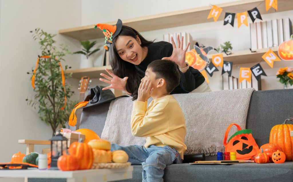 A mother wearing a witch hat playfully "haunts" her son, who is covering his mouth and looking surprised on a couch. The room is decorated for Halloween with banners that say "Happy Halloween" and pumpkins. ©Worawee Meepian