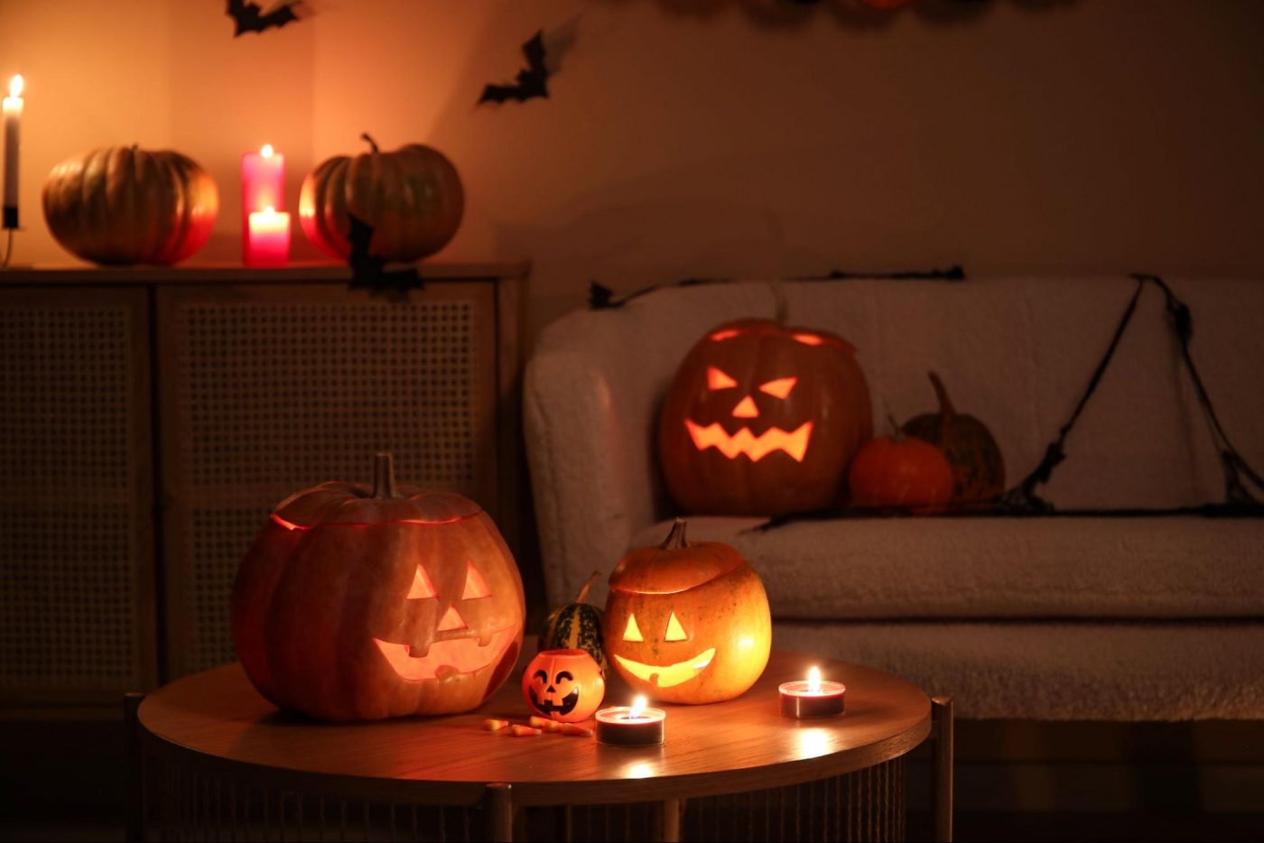 A dark room is lit by the glow of carved jack-o'-lanterns on a coffee table and pumpkins on a nearby dresser. Bat decorations are hanging from the ceiling. ©Pixel-Shot