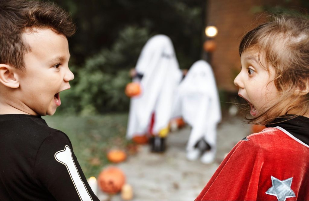A boy and a girl in costume with surprised expressions on their faces look at two blurry, costumed figures in the background. The scene is outdoors on a path decorated with pumpkins. ©Rawpixel.com