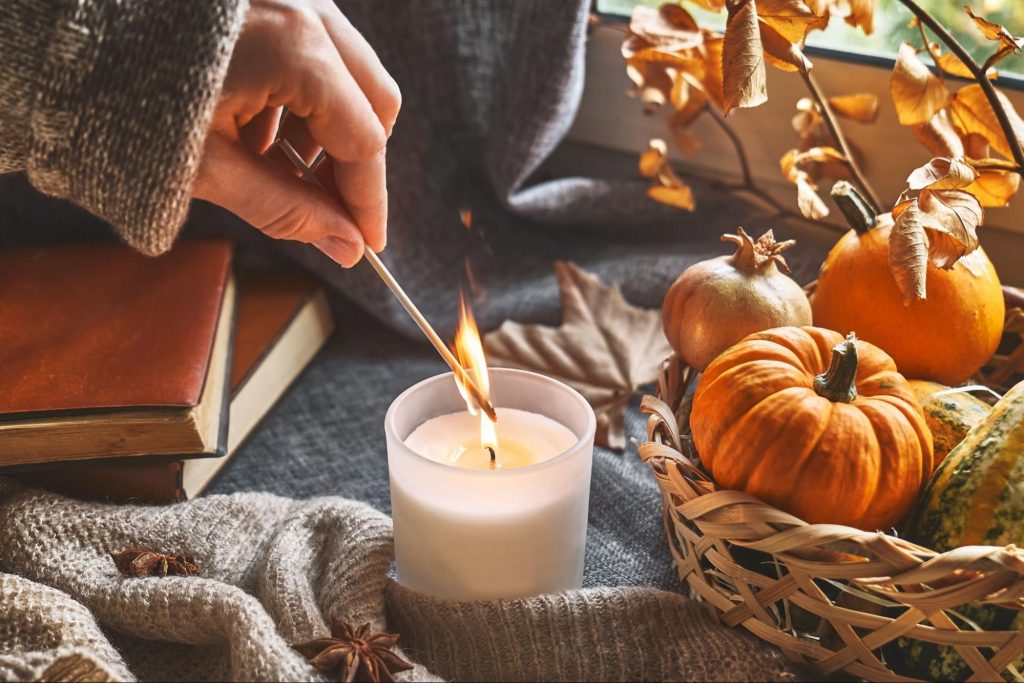 A person's hand lights a white candle with a match. The scene is cozy and autumnal, with a sweater, books, pumpkins, and dried leaves arranged on a gray blanket. ©Caterina Trimarchi