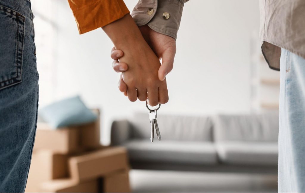 Couple holding hands and house keys inside their new home with moving boxes nearby.
©Prostock-studio